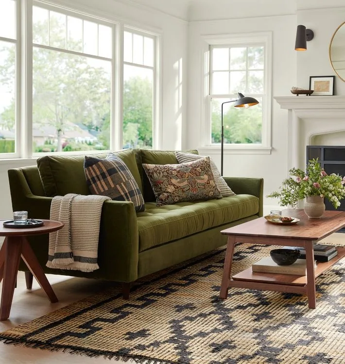 An olive sofa with patterned pillows above a black and beige rug in a white living room.