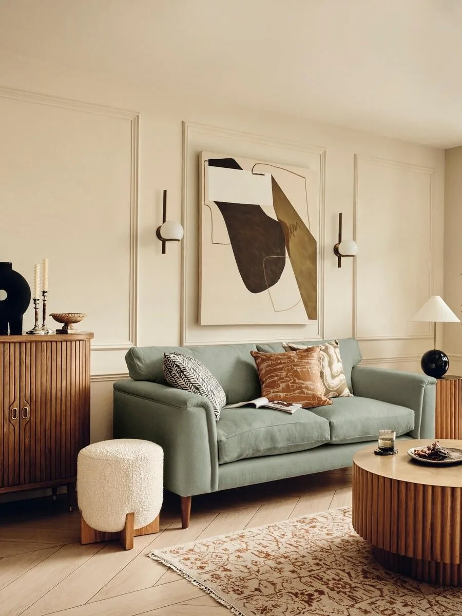 A sage green sofa with ochre and patterned pillows beside cream paneled walls and a patterned rug.