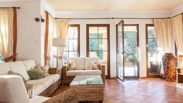 Pale upholstered seats surround a woven table in a room with cream curtains and glazed doors.