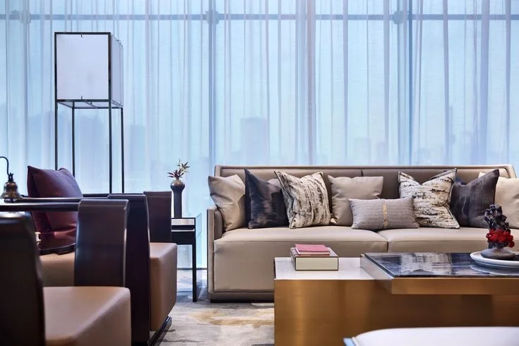 A low beige sofa sits beneath tall windows beside a slim floor lamp and small tables.