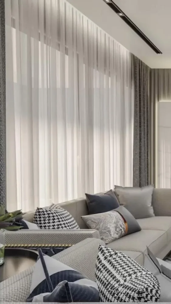 Gray seating with patterned cushions sits beside full height white sheer curtains.